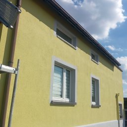 ON BUD Michał Zejer - Żółta elewacja domu z białymi obramowaniami okien i szarą podmurówką. Widoczny fragment rusztowania i błękitne niebo z chmurami. Dom po tynkowaniu.