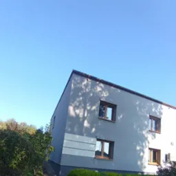 Dwukondygnacyjny dom z szarą elewacją i drewnianymi ramami okiennymi w słoneczny dzień. Na ścianie widoczne cienie drzew. Budynek otoczony zielenią.