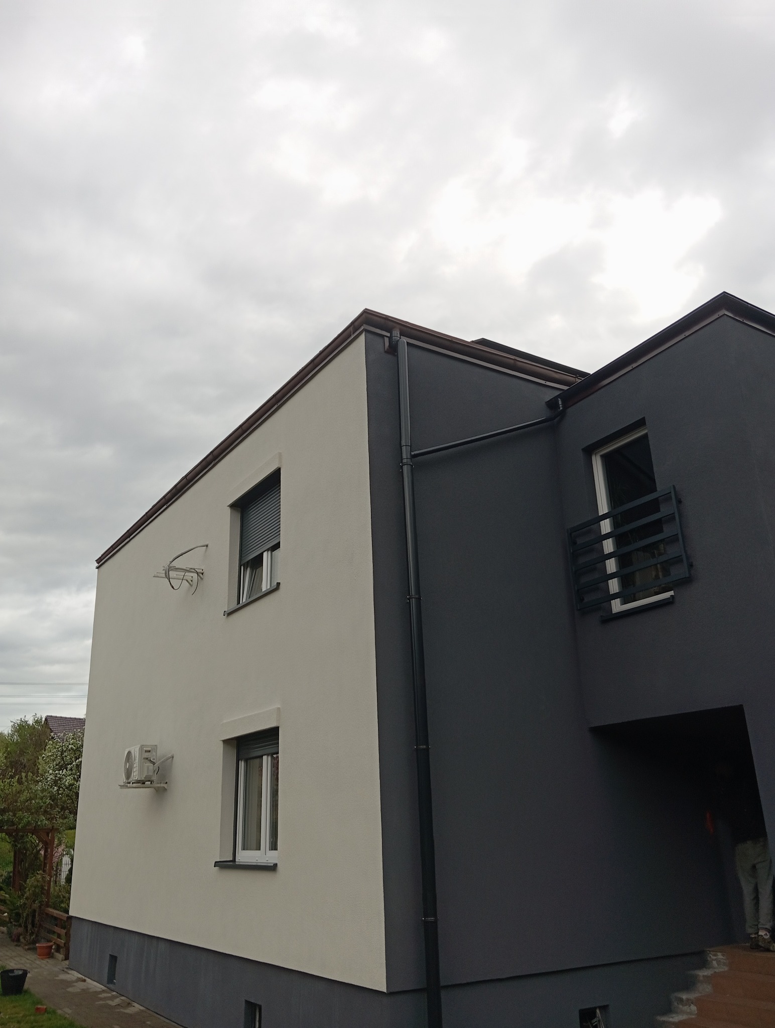 Dwukolorowa elewacja domu z balkonem, jasna ściana z klimatyzatorem i roletami, ciemna z balustradą, widok na pochmurne niebo.