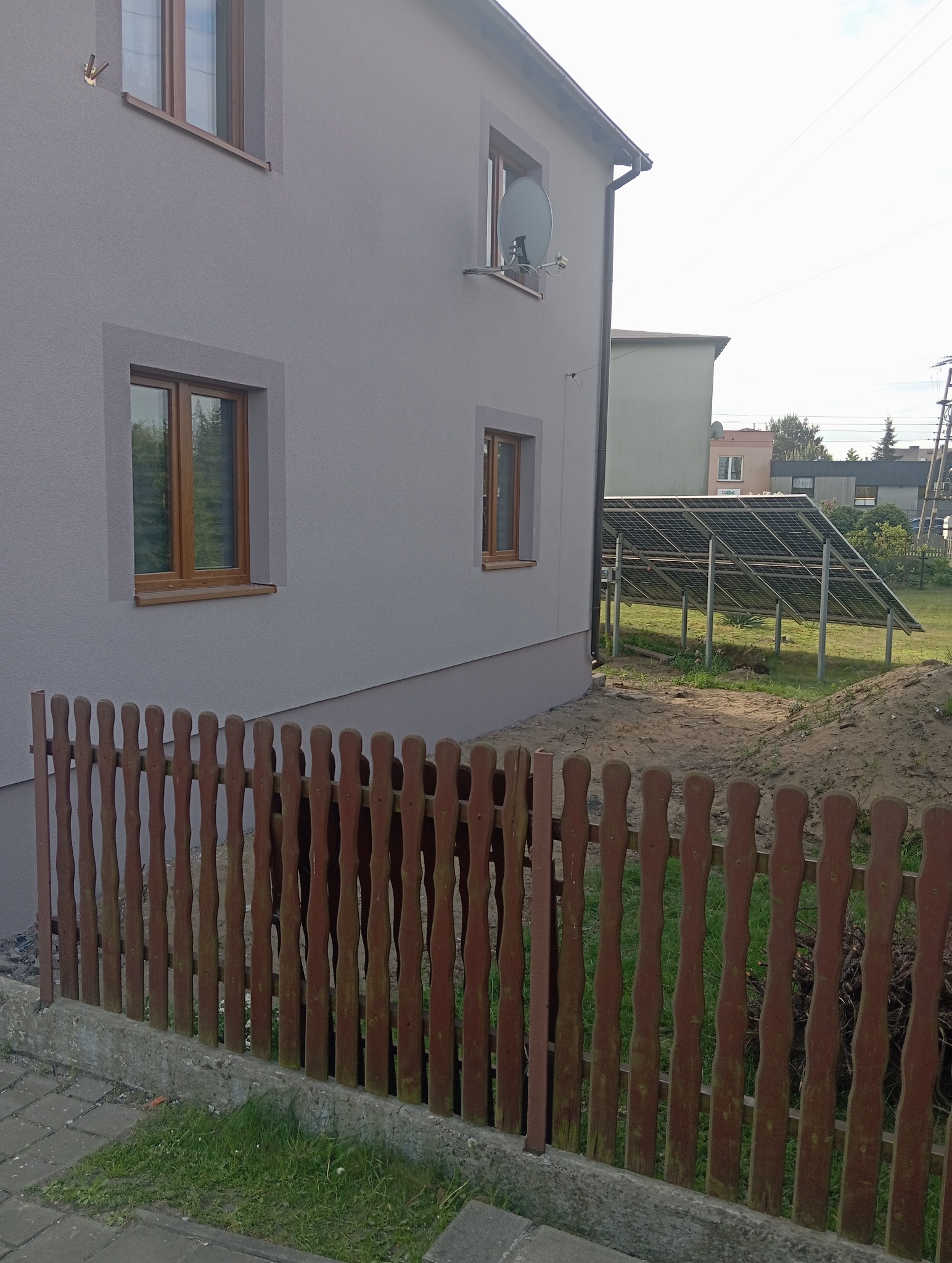 Dom po renowacji elewacji, widoczne okna z brązowymi ramami, elewacja w odcieniach szarości. W tle panele słoneczne i drewniany płot.