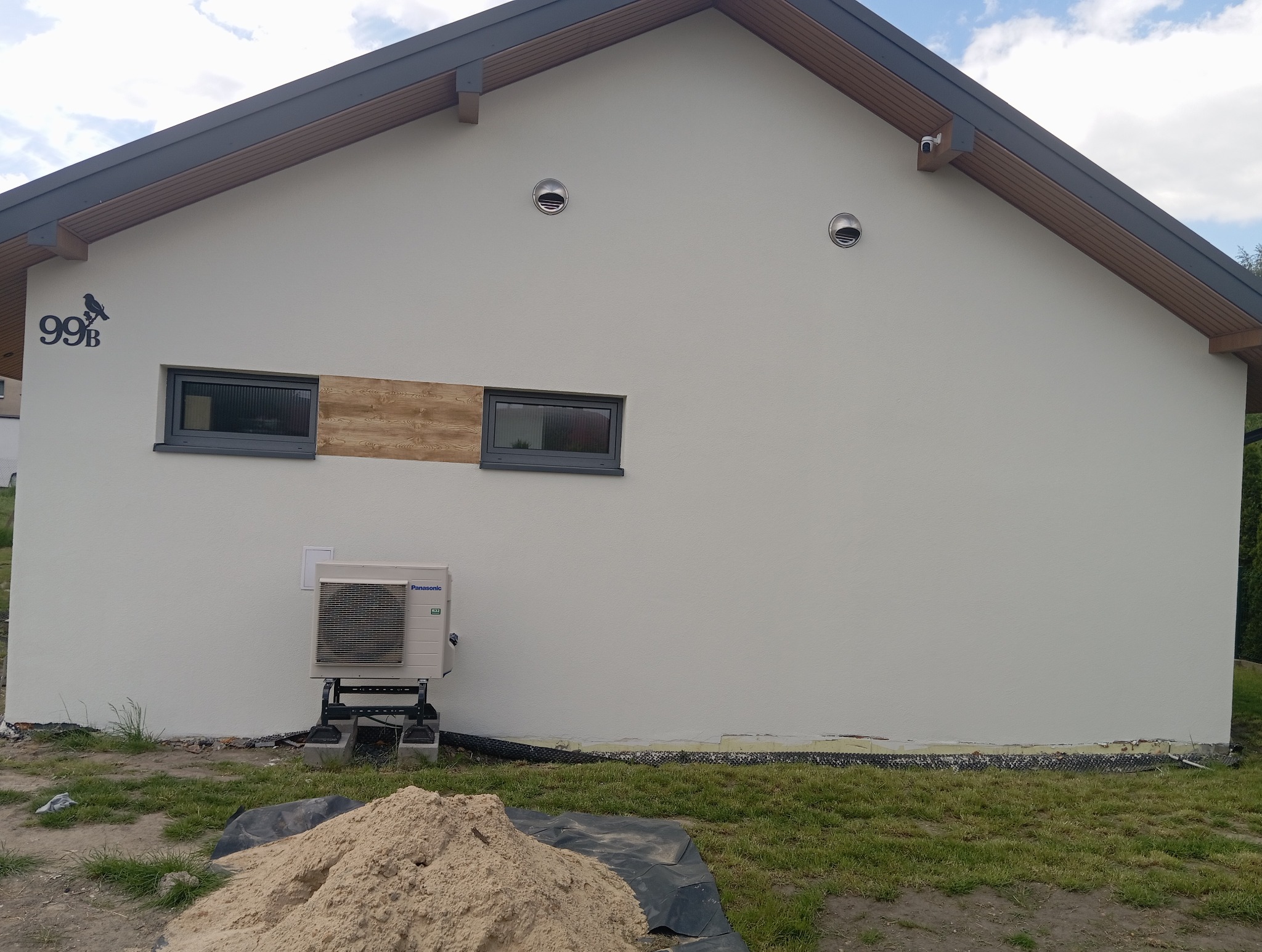Jasna elewacja domu z numerem 99B i drewnianym akcentem obok okna. Widoczny klimatyzator Panasonic i fragment trawnika z piaskiem.