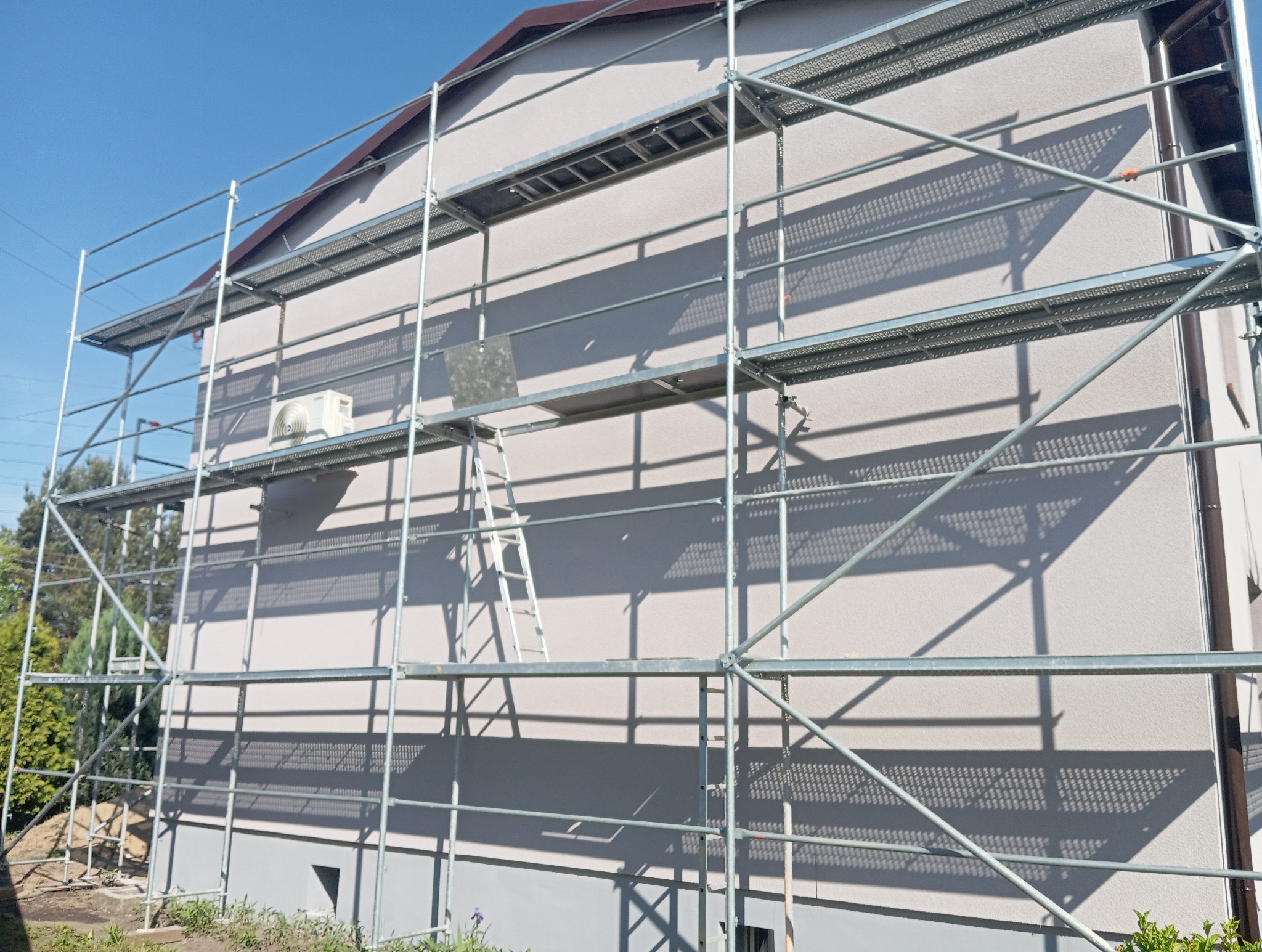 Dom jednorodzinny w trakcie prac elewacyjnych, otoczony rusztowaniem. Widoczny klimatyzator na ścianie i drabina oparta o rusztowanie. Słoneczny dzień.