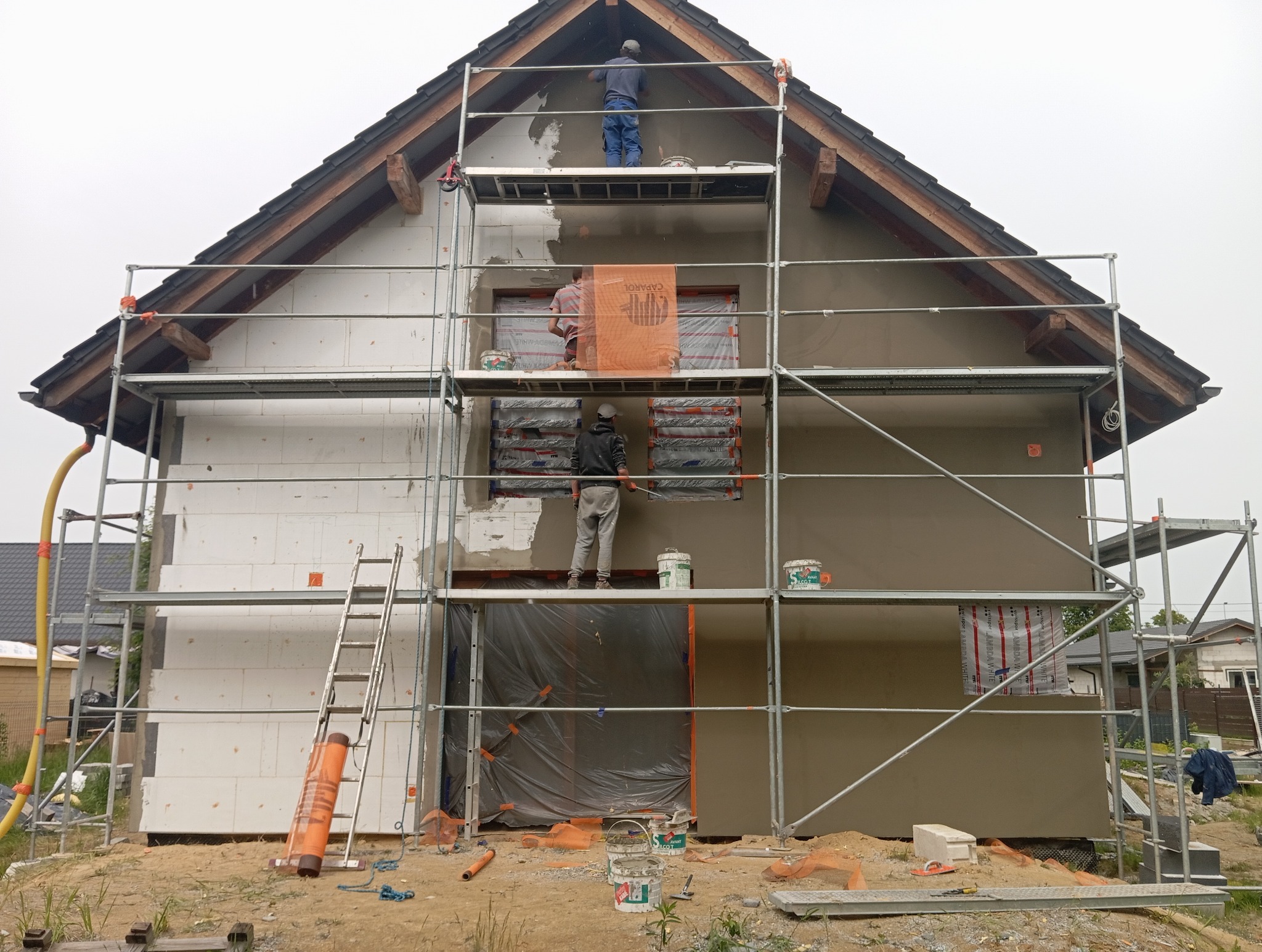 Dom w trakcie ocieplania: jedna ściana z izolacją, druga z nałożonym tynkiem. Ekipa na rusztowaniu wykonuje prace wykończeniowe elewacji.