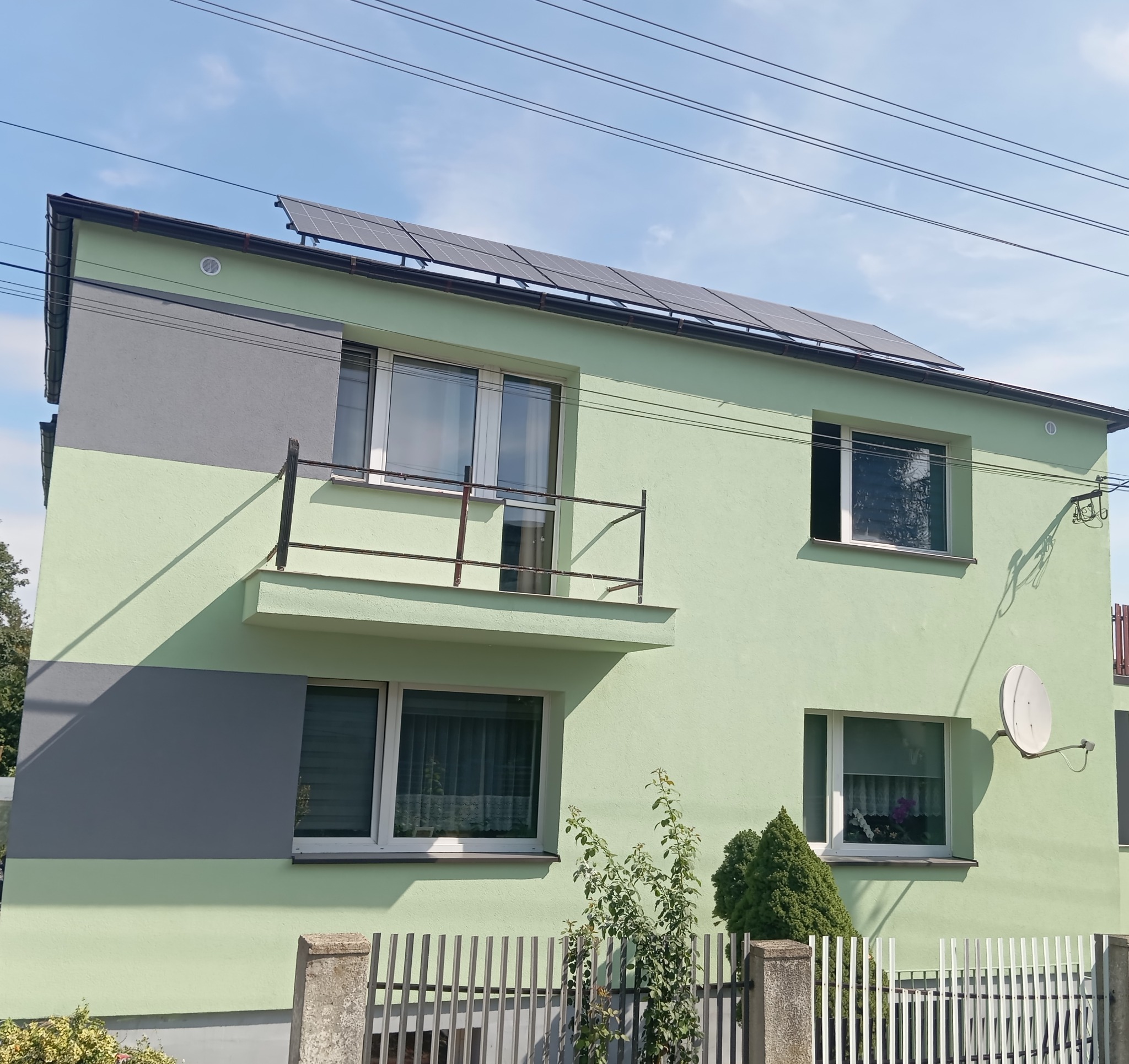 Dom z balkonem po malowaniu elewacji na jasnozielono i szaro, z panelami słonecznymi na dachu. Widoczna antena satelitarna i ogrodzenie.