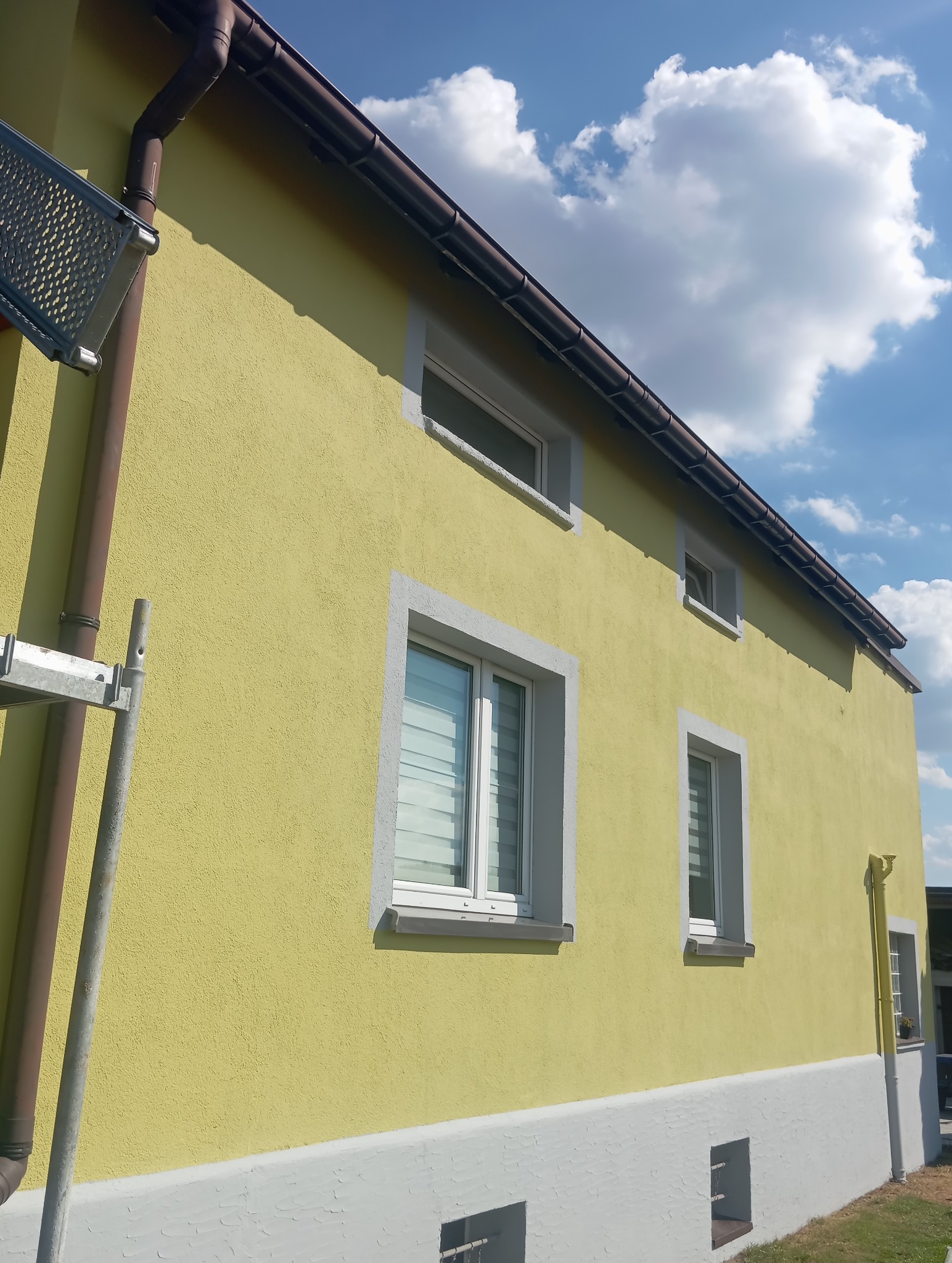 Żółta elewacja domu z białymi obramowaniami okien i szarą podmurówką. Widoczny fragment rusztowania i błękitne niebo z chmurami. Dom po tynkowaniu.
