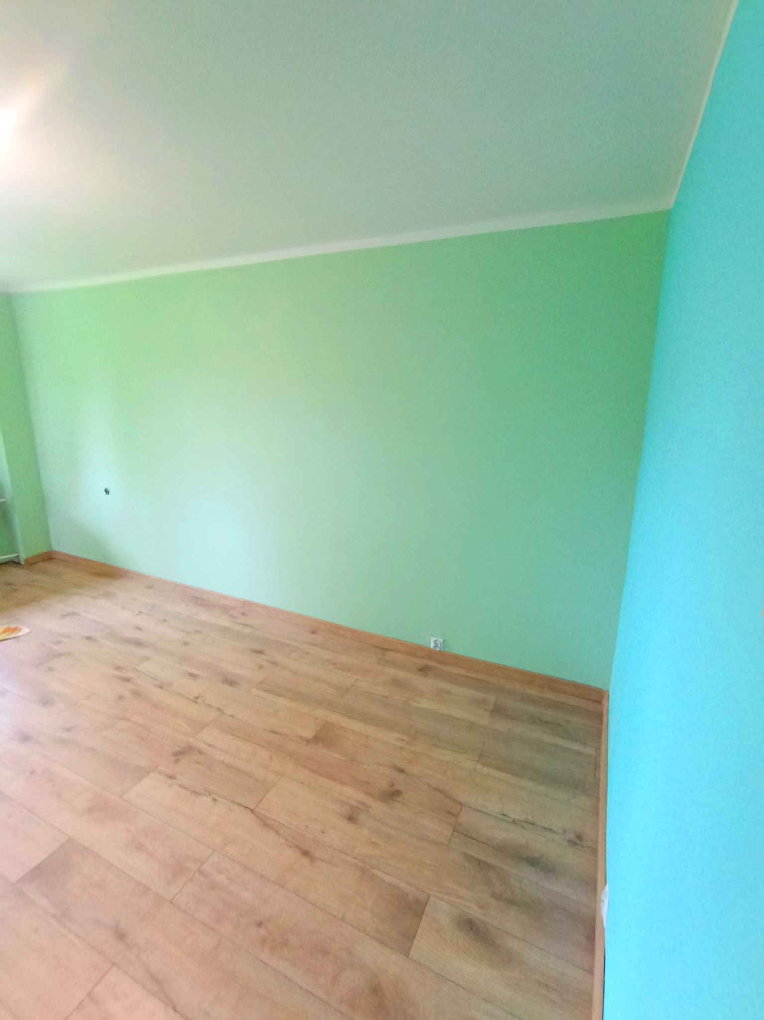 Pomieszczenie po odświeżeniu: ściany pomalowane na zielono i błękitno, drewniana podłoga. Widok na odnowione wnętrze z perspektywy podłogi.