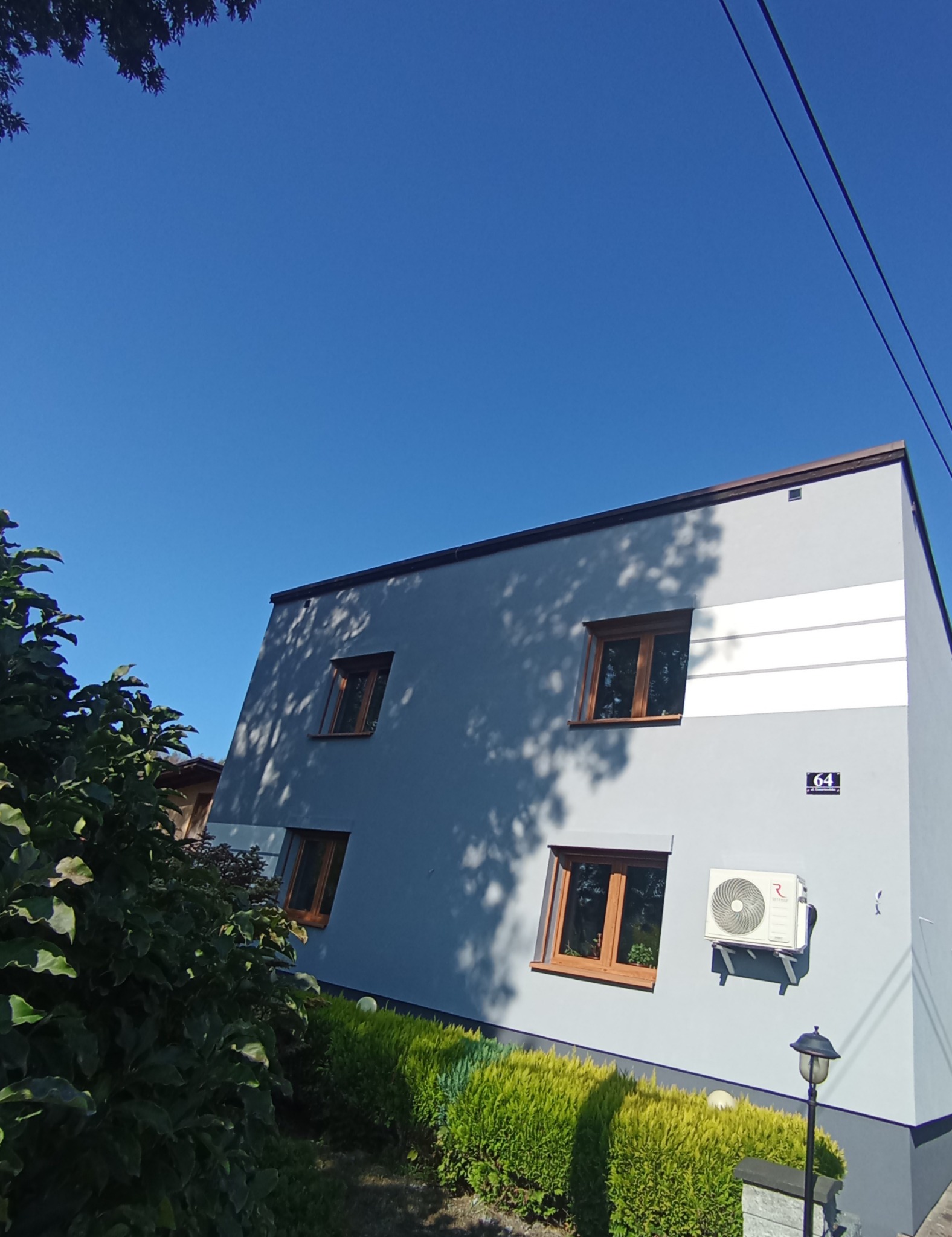 Jasnoszara elewacja domu z drewnianymi oknami w słoneczny dzień. Widoczny klimatyzator, numer domu i cień drzew. Ujęcie z dołu, zieleń w tle.