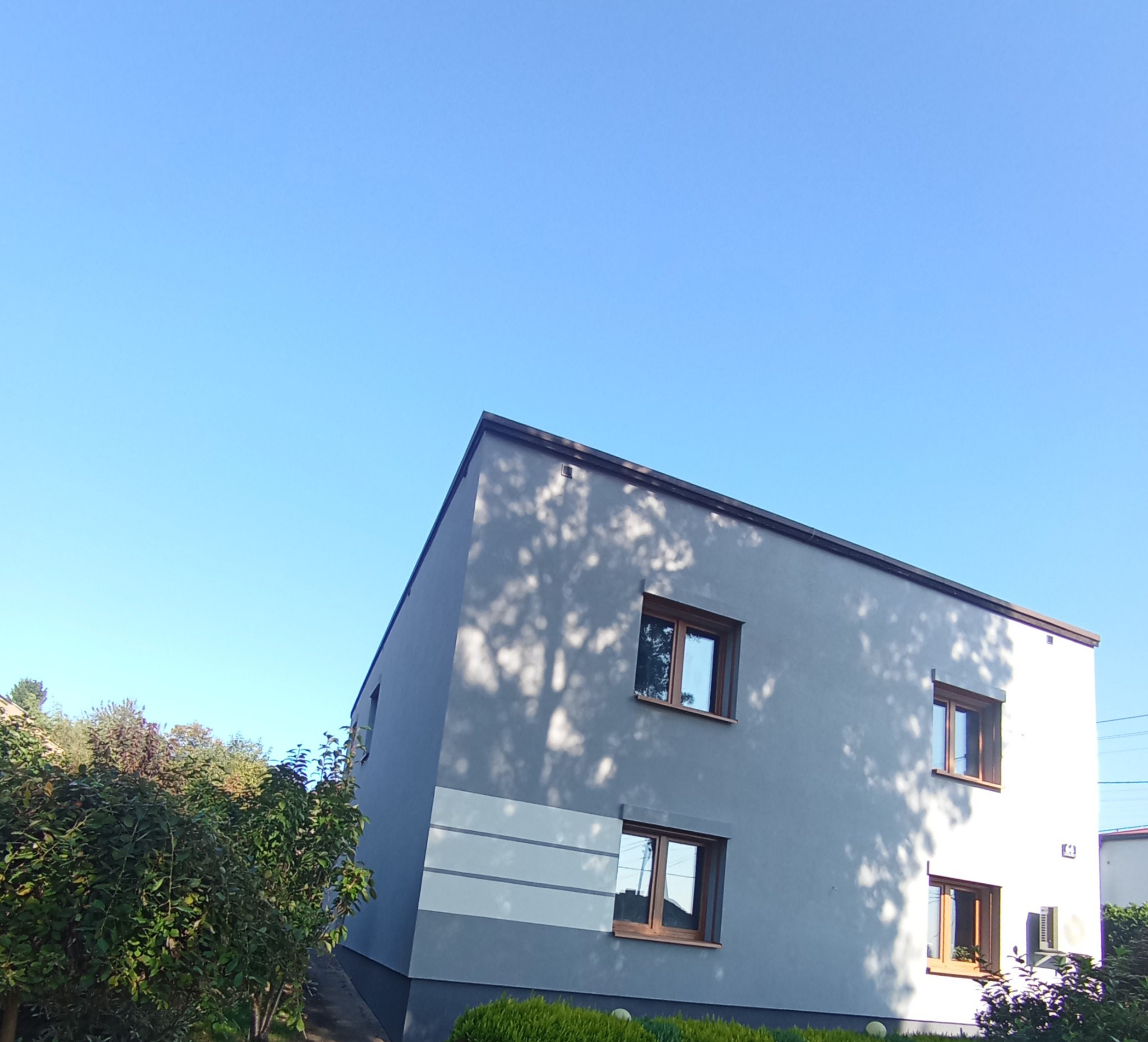 Dwukondygnacyjny dom z szarą elewacją i drewnianymi ramami okiennymi w słoneczny dzień. Na ścianie widoczne cienie drzew. Budynek otoczony zielenią.