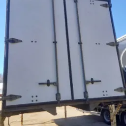 Białe, dwuskrzydłowe drzwi naczepy po renowacji, widoczne metalowe zawiasy i zamknięcia. Drzwi zamontowane na naczepie, w tle błękitne niebo.