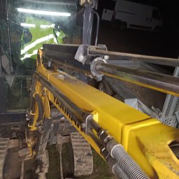 Mobilne Usługi Spawalnicze - Adam Słomian - Żółte ramię minikoparki Yanmar z widocznymi przewodami hydraulicznymi i gąsienicami, nocne oświetlenie, serwis w terenie, w tle biały bus.