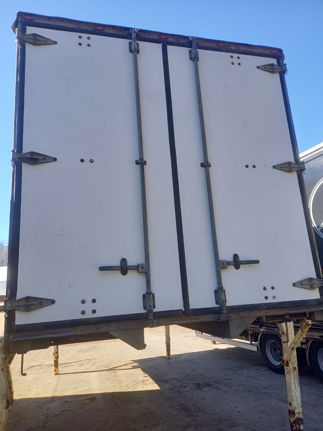 Białe, dwuskrzydłowe drzwi naczepy po renowacji, widoczne metalowe zawiasy i zamknięcia. Drzwi zamontowane na naczepie, w tle błękitne niebo.