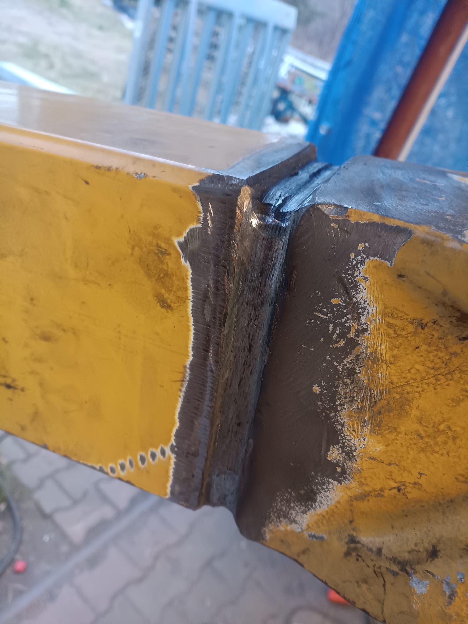 Żółty element konstrukcyjny po spawaniu, widoczne ślady naprawy pęknięcia. Szlifowana spoina na metalu, przygotowana do dalszej obróbki. Detal z bliska.
