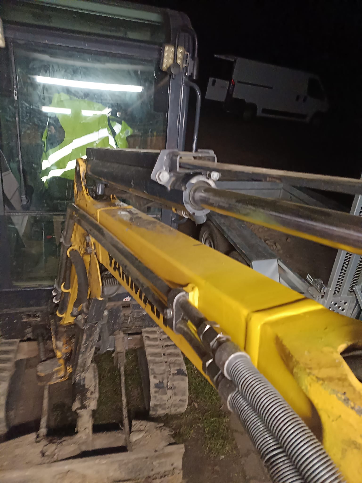 Żółte ramię minikoparki Yanmar z widocznymi przewodami hydraulicznymi i gąsienicami, nocne oświetlenie, serwis w terenie, w tle biały bus.