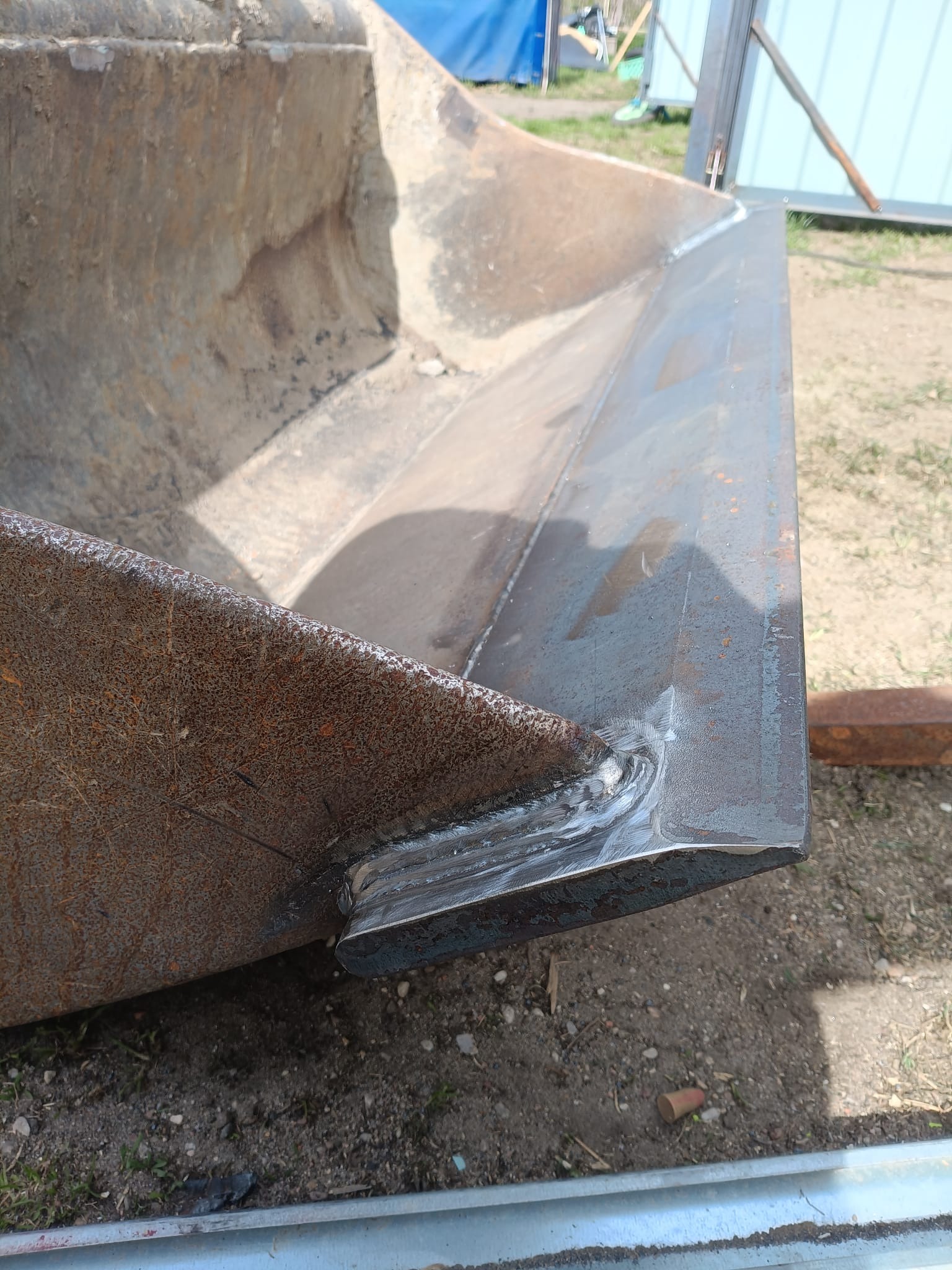 Detal spoiny na krawędzi łyżki skarpowej koparki po naprawie. Widoczne ślady rdzy i ziemi. Tło stanowi grunt i fragment konstrukcji.