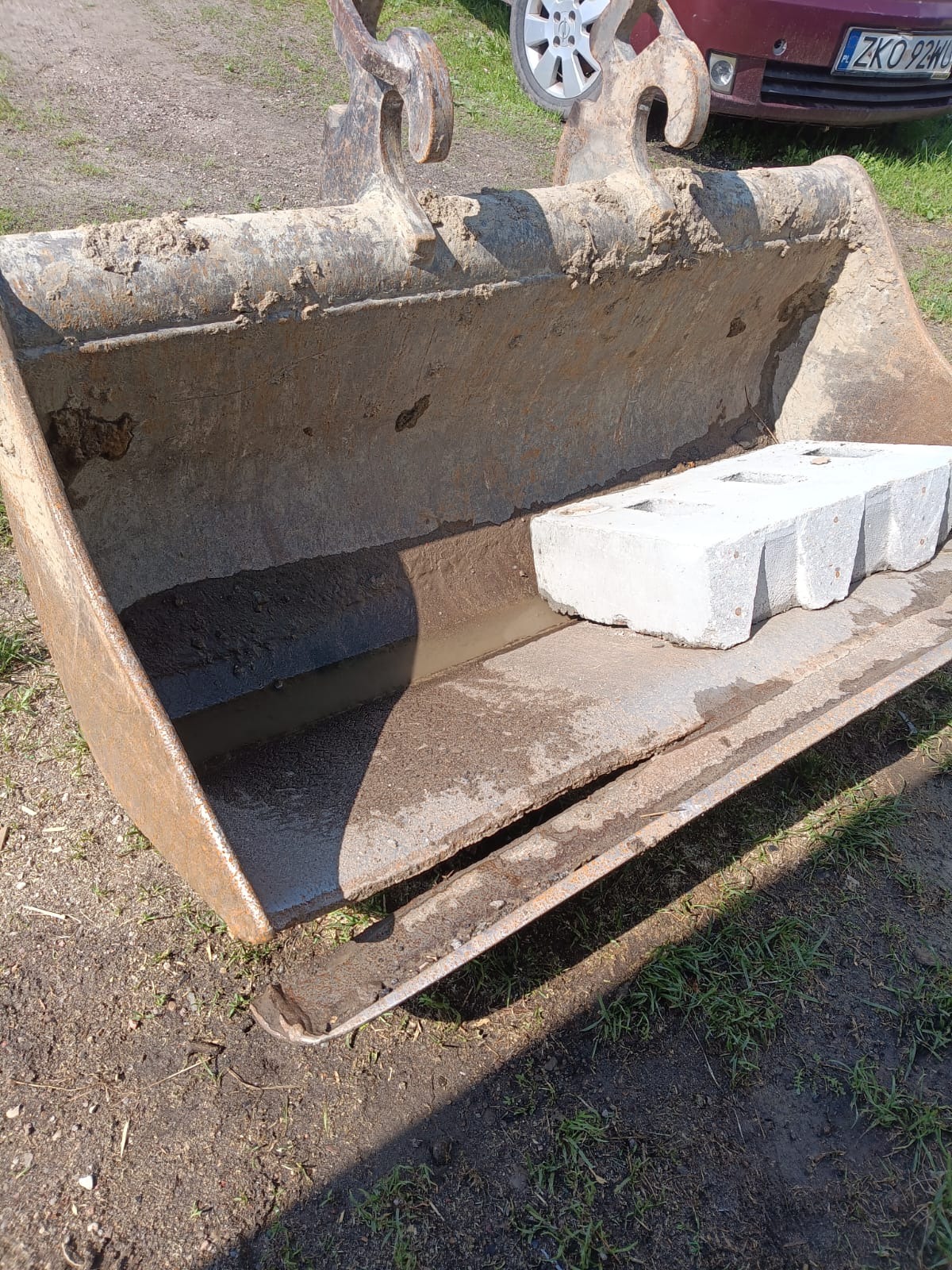 Skarpa łyżki koparki po naprawie spawalniczej, widoczne ślady spawania. Wnętrze łyżki wypełnione wodą i betonowym elementem. Częściowo widoczny samochód osobowy.