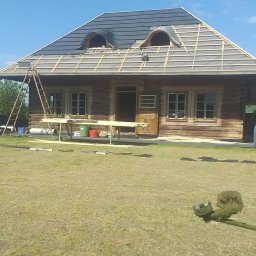 Adrian Suciu - Drewniany dom z dachem w trakcie krycia czarną dachówką, widoczne okna dachowe, rusztowanie, trawa i drabina. Częściowo pokryty dach.