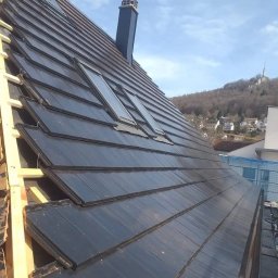 Adrian Suciu - Nowy, grafitowy dach z trzema oknami dachowymi, częściowo pokryty, z widoczną konstrukcją drewnianą i kominem. W tle zabudowa miejska i zalesione wzgórze.