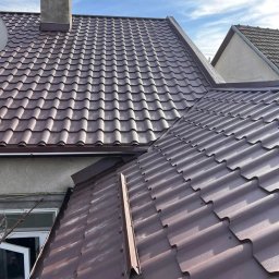 Adrian Suciu - Brązowy dach z blachy trapezowej, widok z góry. Kominek w tle, fragment okna i rynny widoczne w lewym dolnym rogu. Błękitne niebo w tle.