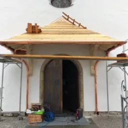 Kościół w trakcie renowacji dachu z drewnianą konstrukcją, miedzianymi rynnami i rusztowaniem. Częściowo otwarte, drewniane drzwi wejściowe.