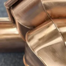 Miedziane, ręcznie robione elementy orynnowania, z precyzyjnymi łączeniami, ukazane z bliska na szarym tle. Detal obr&oacute;bki blacharskiej z połyskiem.