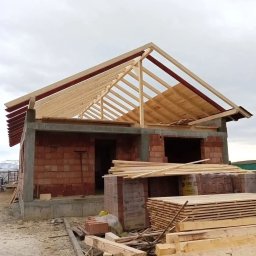 Adrian Suciu - Surowy stan: budowa domu z odsłoniętą więźbą dachową z drewna, częściowo pokrytą deskami, na tle pochmurnego nieba. Widoczne materiały budowlane.