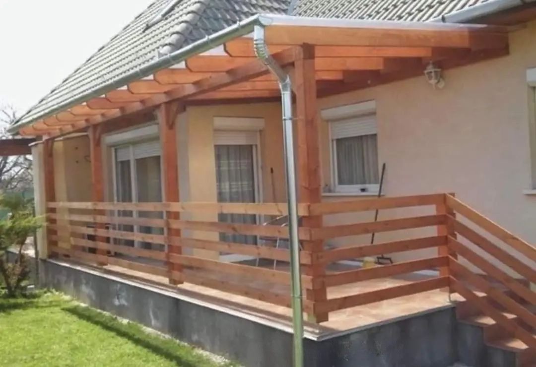 Drewniana pergola z rynnami przy domu z beżową elewacją i szarym dachem. Widoczne drewniane barierki i schody. Elementy konstrukcyjne wykonane z jasnego drewna.