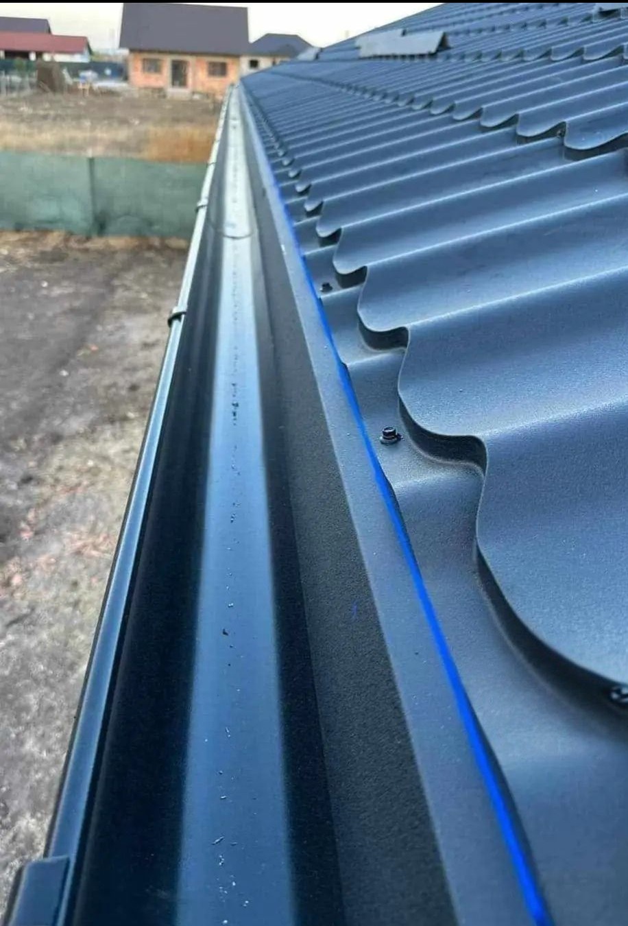 Szczegółowe ujęcie nowej, grafitowej rynny zamontowanej przy dachu z blachy trapezowej. Widoczne mocowania i linia dachu w tle.