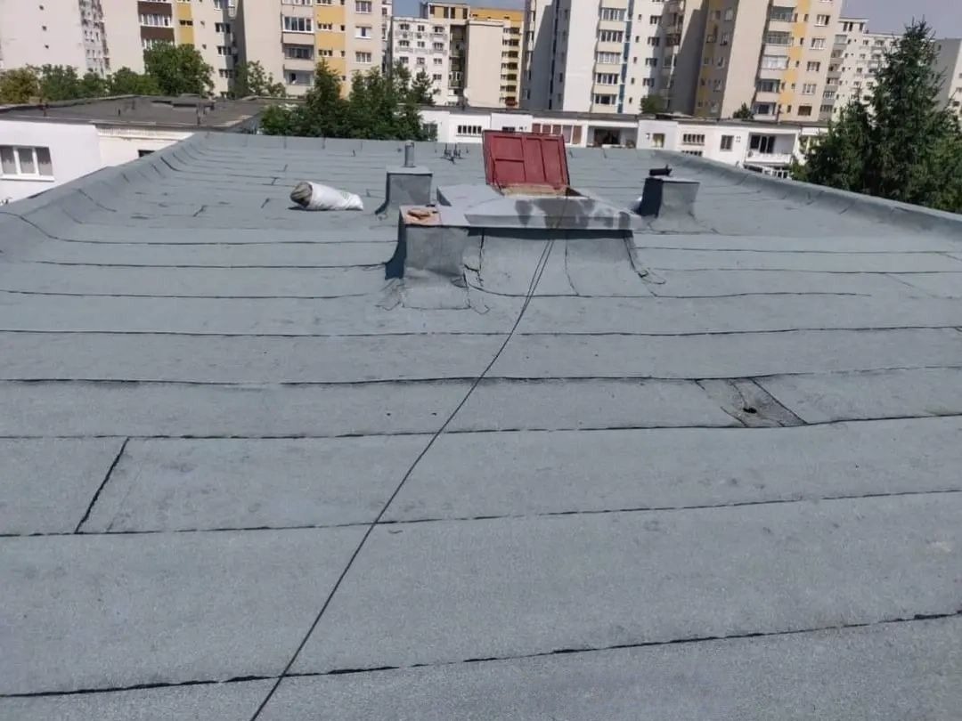 Szary, płaski dach pokryty papą na tle miejskiego krajobrazu z blokami. Widoczne detale: komin i otwarty właz dachowy z czerwoną klapą.