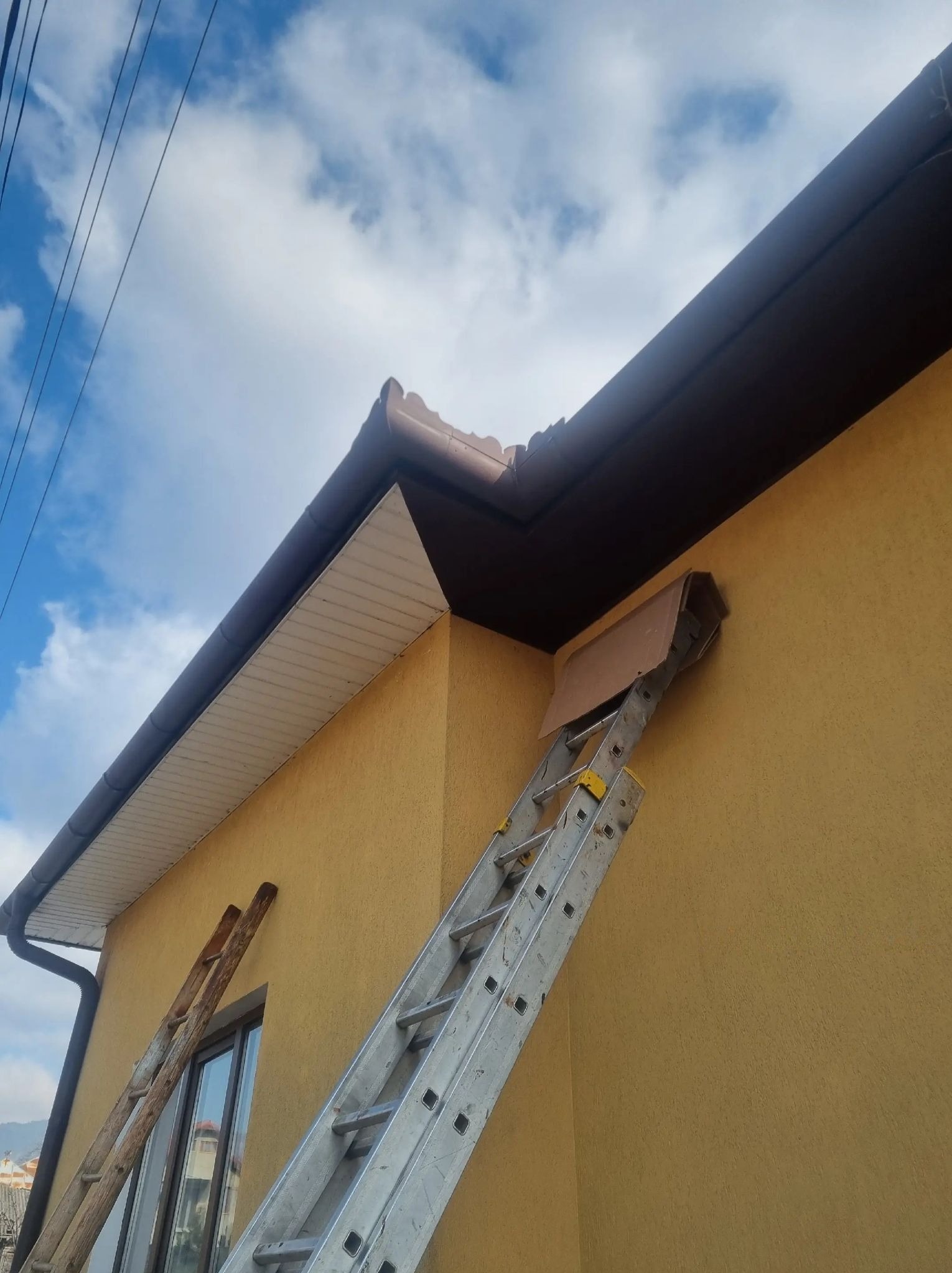 Żółta ściana domu z brązową rynną, dwie drabiny oparte o ścianę, błękitne niebo z chmurami w tle. Ujęcie z dołu.