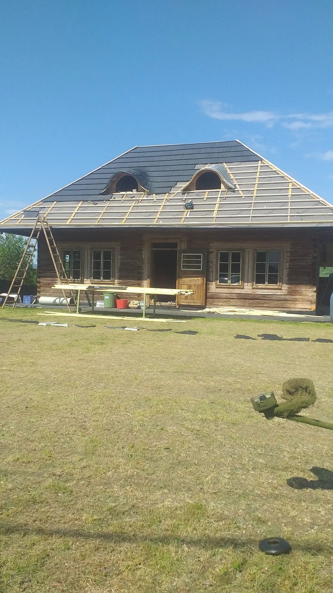 Drewniany dom z dachem w trakcie krycia czarną dachówką, widoczne okna dachowe, rusztowanie, trawa i drabina. Częściowo pokryty dach.