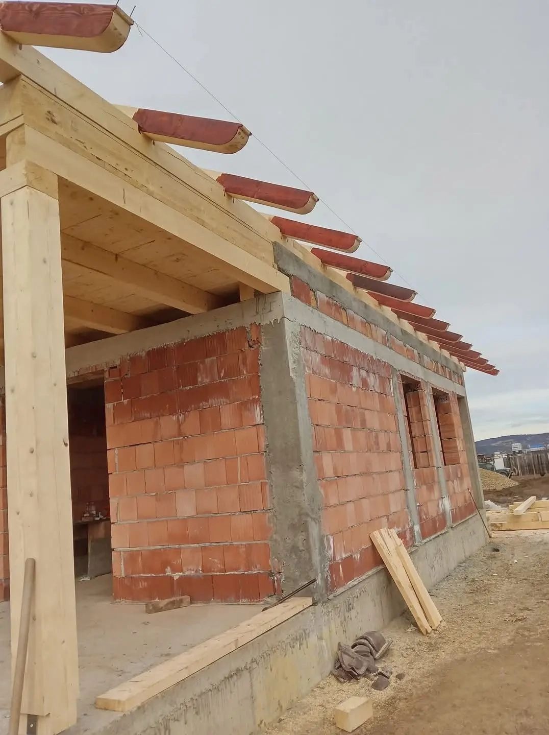 Budowa domu jednorodzinnego w trakcie realizacji: ściany z czerwonej cegły, drewniana więźba dachowa z elementami mocującymi rynny, widok z zewnątrz.