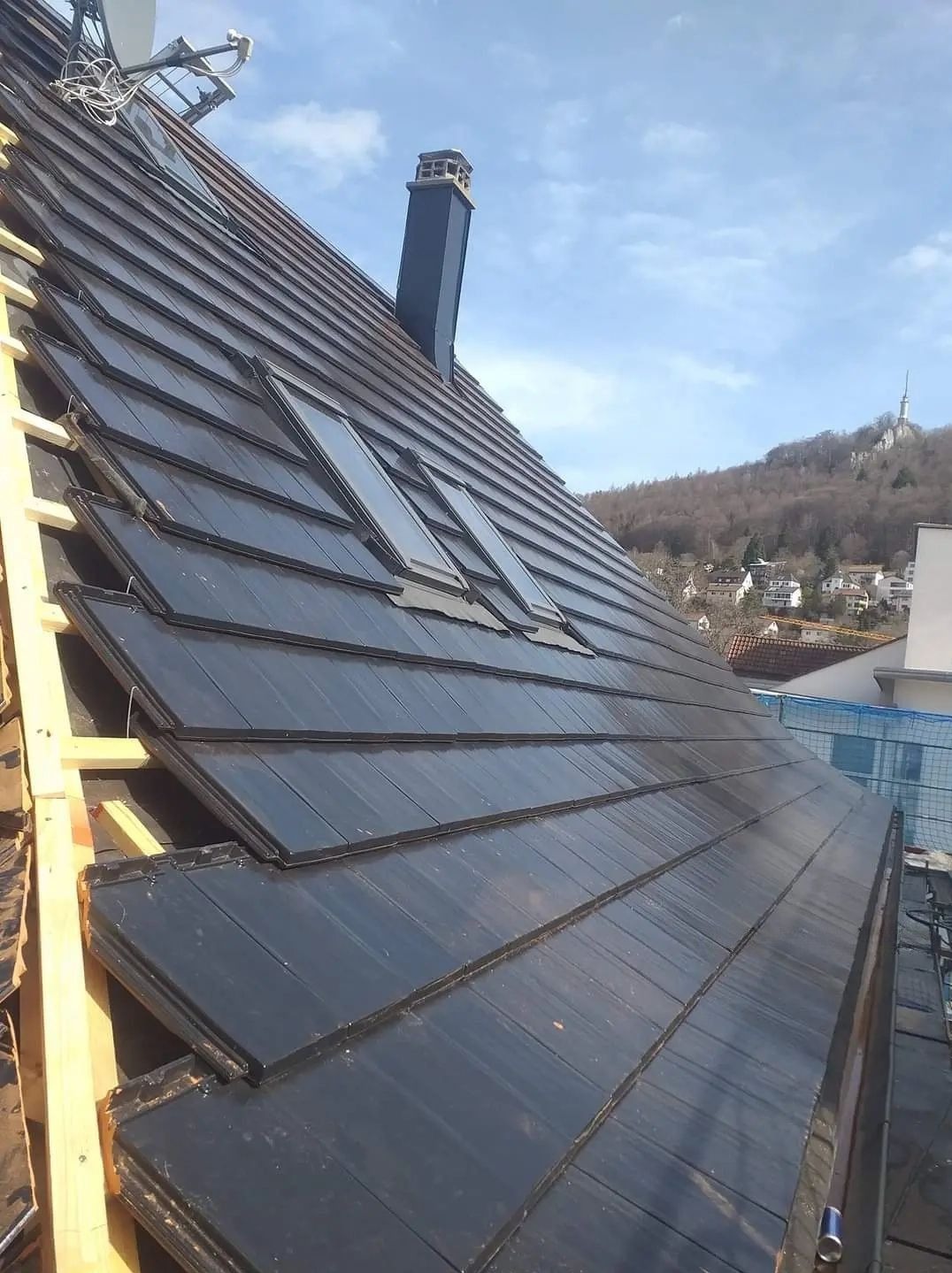 Nowy, grafitowy dach z trzema oknami dachowymi, częściowo pokryty, z widoczną konstrukcją drewnianą i kominem. W tle zabudowa miejska i zalesione wzgórze.