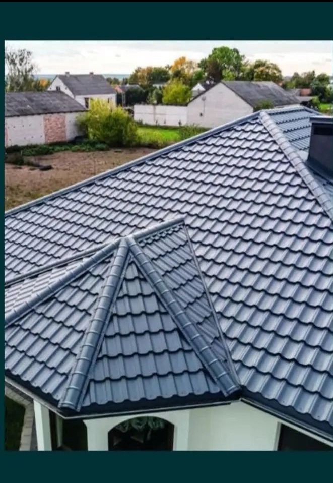 Szary dach z blachodachówki modułowej na domu jednorodzinnym, widok z góry, na tle zabudowań i zieleni w słoneczny dzień.