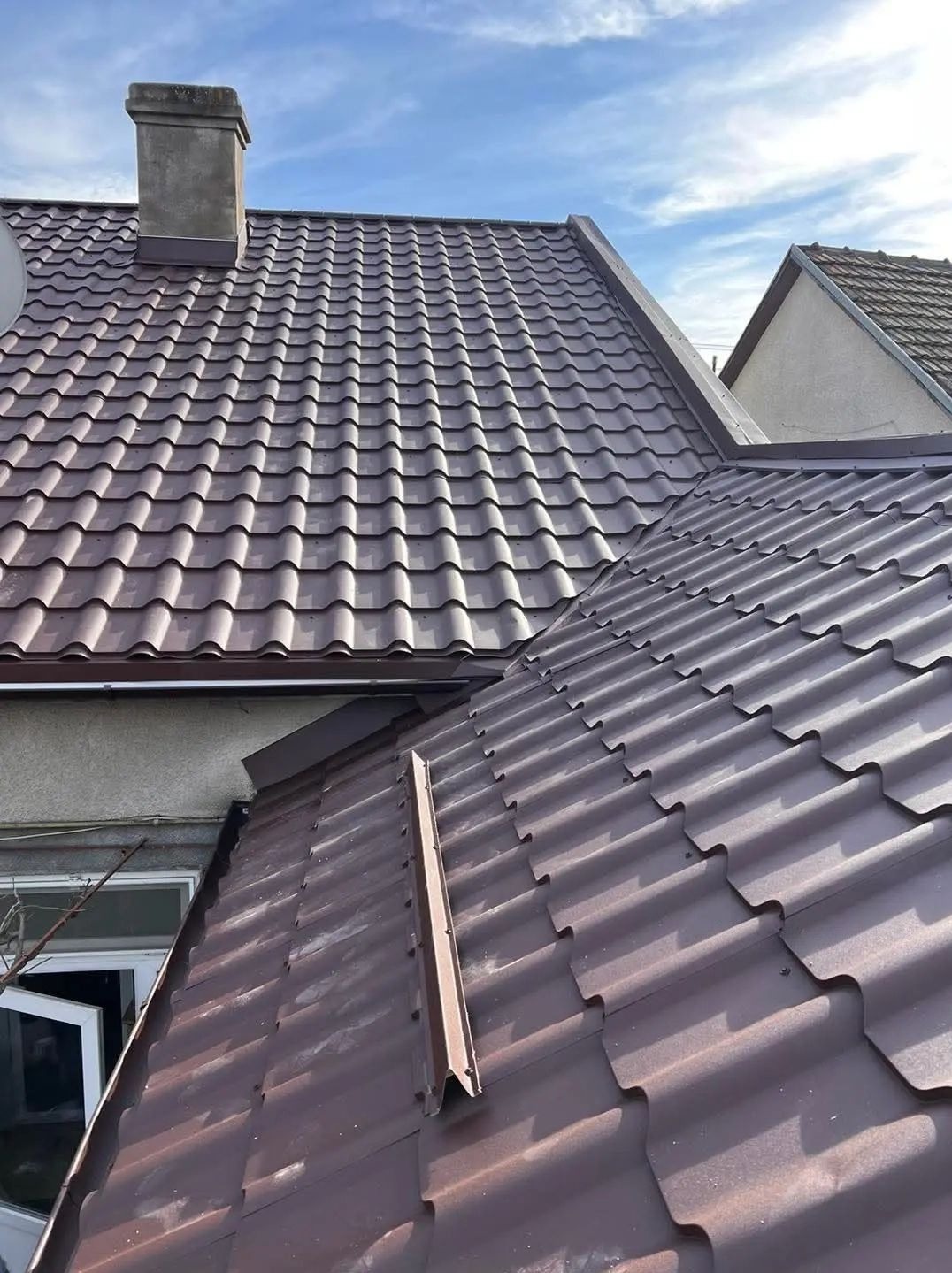 Brązowy dach z blachy trapezowej, widok z góry. Kominek w tle, fragment okna i rynny widoczne w lewym dolnym rogu. Błękitne niebo w tle.