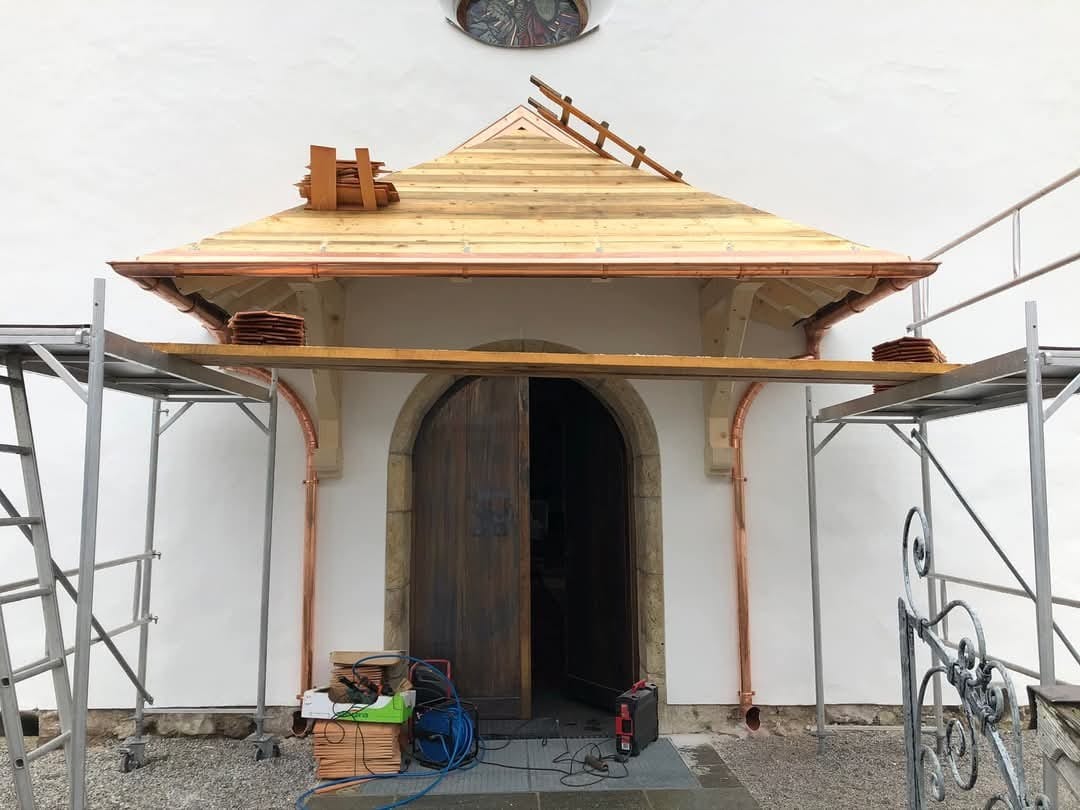 Kościół w trakcie renowacji dachu z drewnianą konstrukcją, miedzianymi rynnami i rusztowaniem. Częściowo otwarte, drewniane drzwi wejściowe.