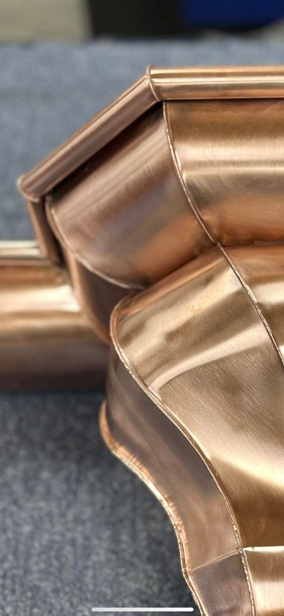 Miedziane, ręcznie robione elementy orynnowania, z precyzyjnymi łączeniami, ukazane z bliska na szarym tle. Detal obróbki blacharskiej z połyskiem.