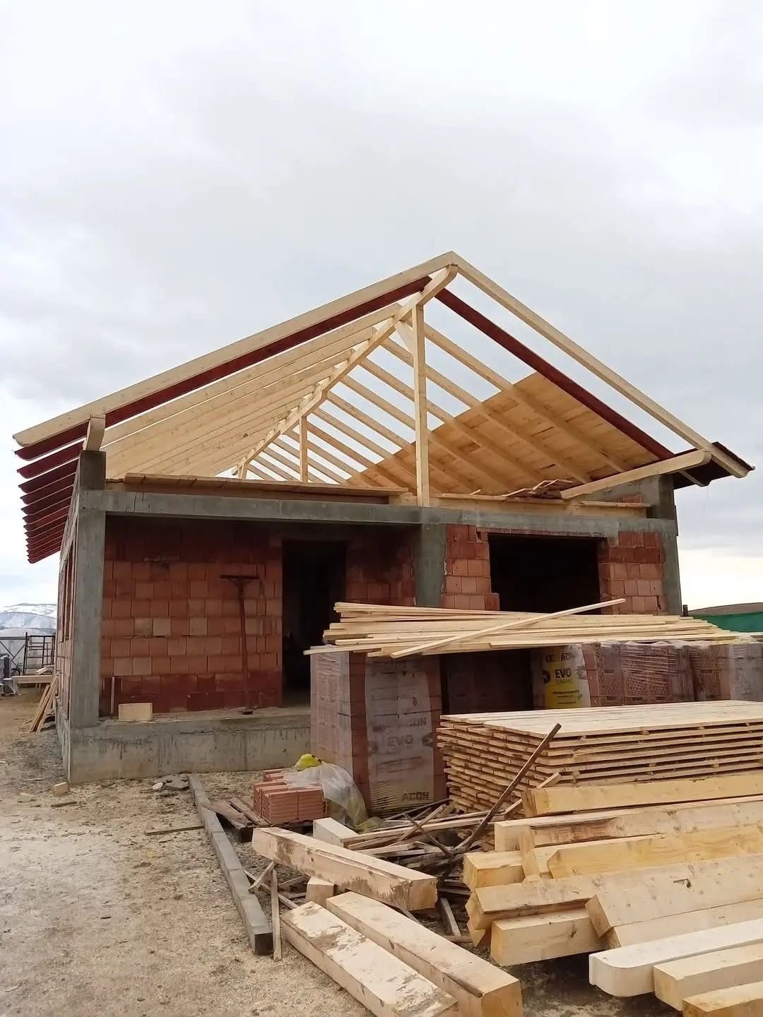 Surowy stan: budowa domu z odsłoniętą więźbą dachową z drewna, częściowo pokrytą deskami, na tle pochmurnego nieba. Widoczne materiały budowlane.