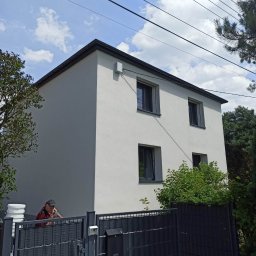 RTJ-BUD - Dwupiętrowy dom z jasną elewacją, widoczny zza czarnego ogrodzenia. Na pierwszym planie fragment samochodu i osoba w czerwonej koszuli. Błękitne niebo z chmurami w tle.