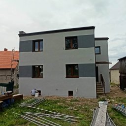 RTJ-BUD - Dwukondygnacyjny dom po tynkowaniu elewacji w odcieniach szarości, z widocznymi oknami z brązowymi ramami. Na trawie leżą elementy rusztowania, a obok pracuje osoba.