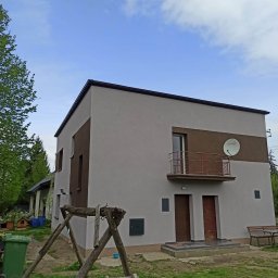 RTJ-BUD - Dwukondygnacyjny dom z balkonem i anteną satelitarną, pomalowany w odcieniach szarości i brązu, otoczony zielenią. Widoczne elementy placu zabaw.