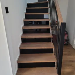 Nowoczesne schody drewniane z czarnymi podstopnicami i metalową balustradą. Widok z dołu, ukazujący detale wykończenia i naturalny odcień drewna.