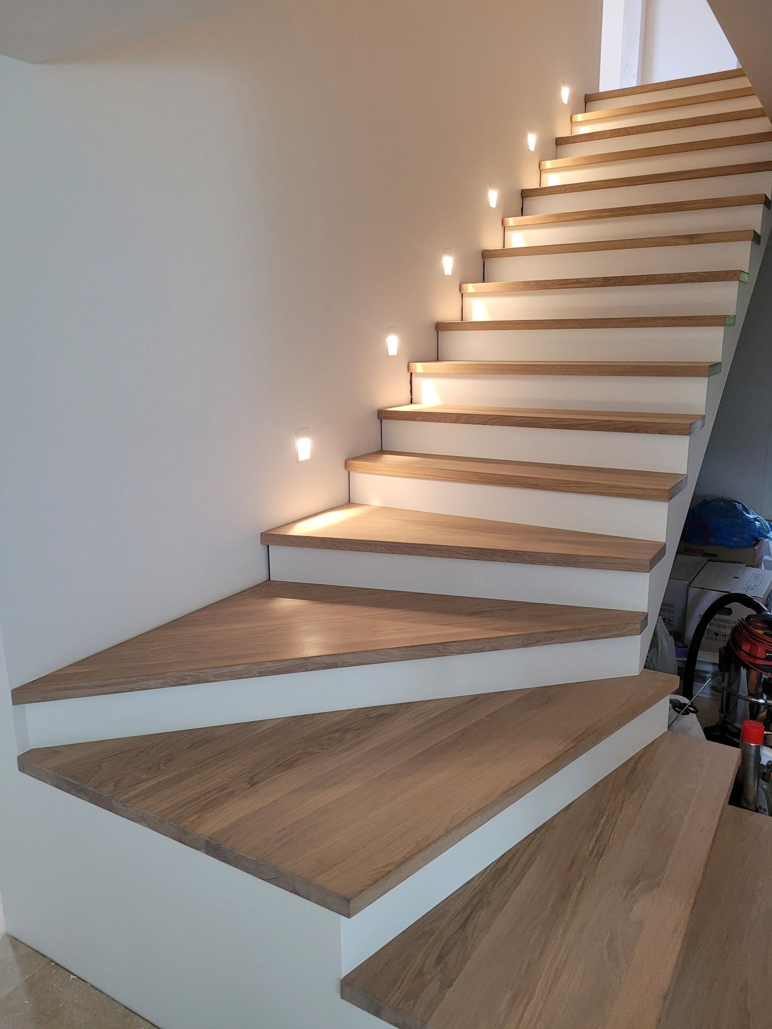 Nowoczesne schody z drewnianymi stopniami i białymi podstopnicami, oświetlone punktowymi lampkami w ścianie. Widok z dołu, ukazujący trójkątny kształt pierwszego stopnia.