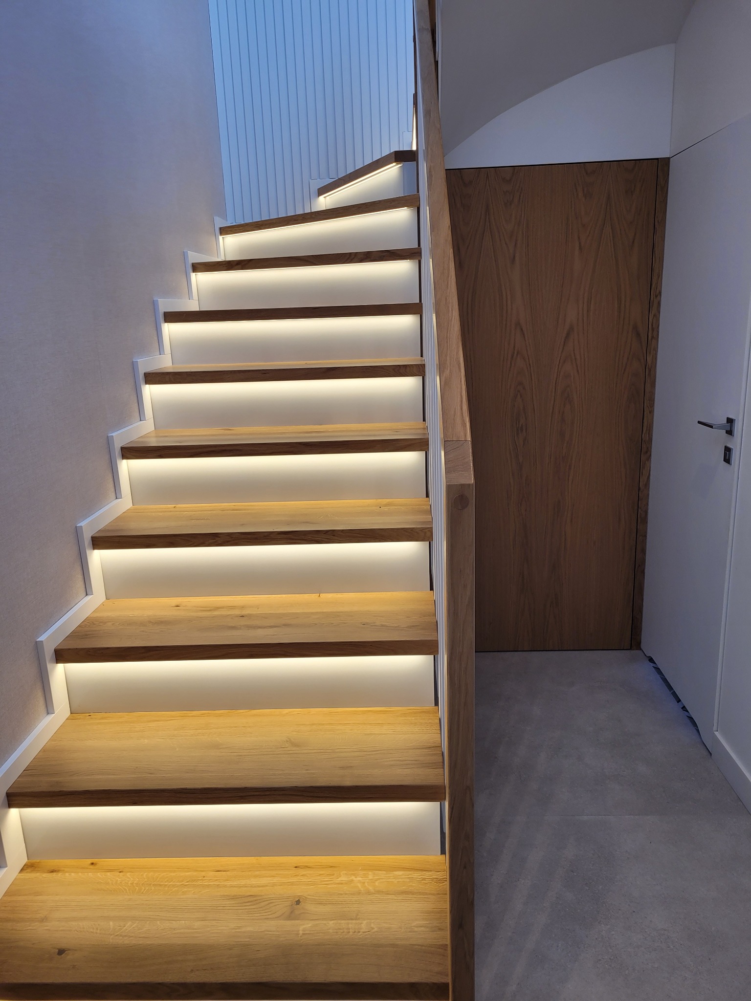 Nowoczesne schody z drewna dębowego z białymi podstopnicami i subtelnym oświetleniem LED, prowadzące do góry. Widoczna drewniana balustrada i drzwi w tle.