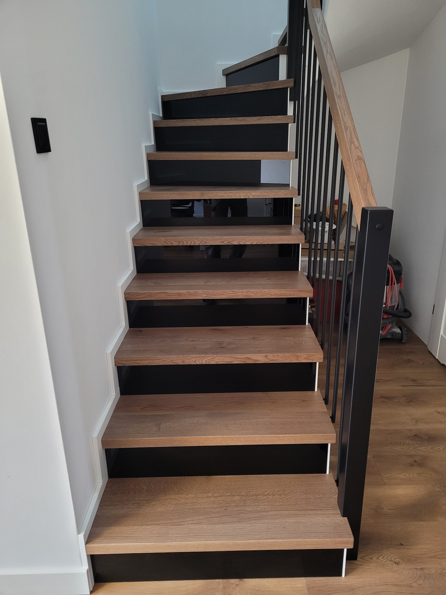 Nowoczesne schody drewniane z czarnymi podstopnicami i metalową balustradą. Widok z dołu, ukazujący detale wykończenia i naturalny odcień drewna.