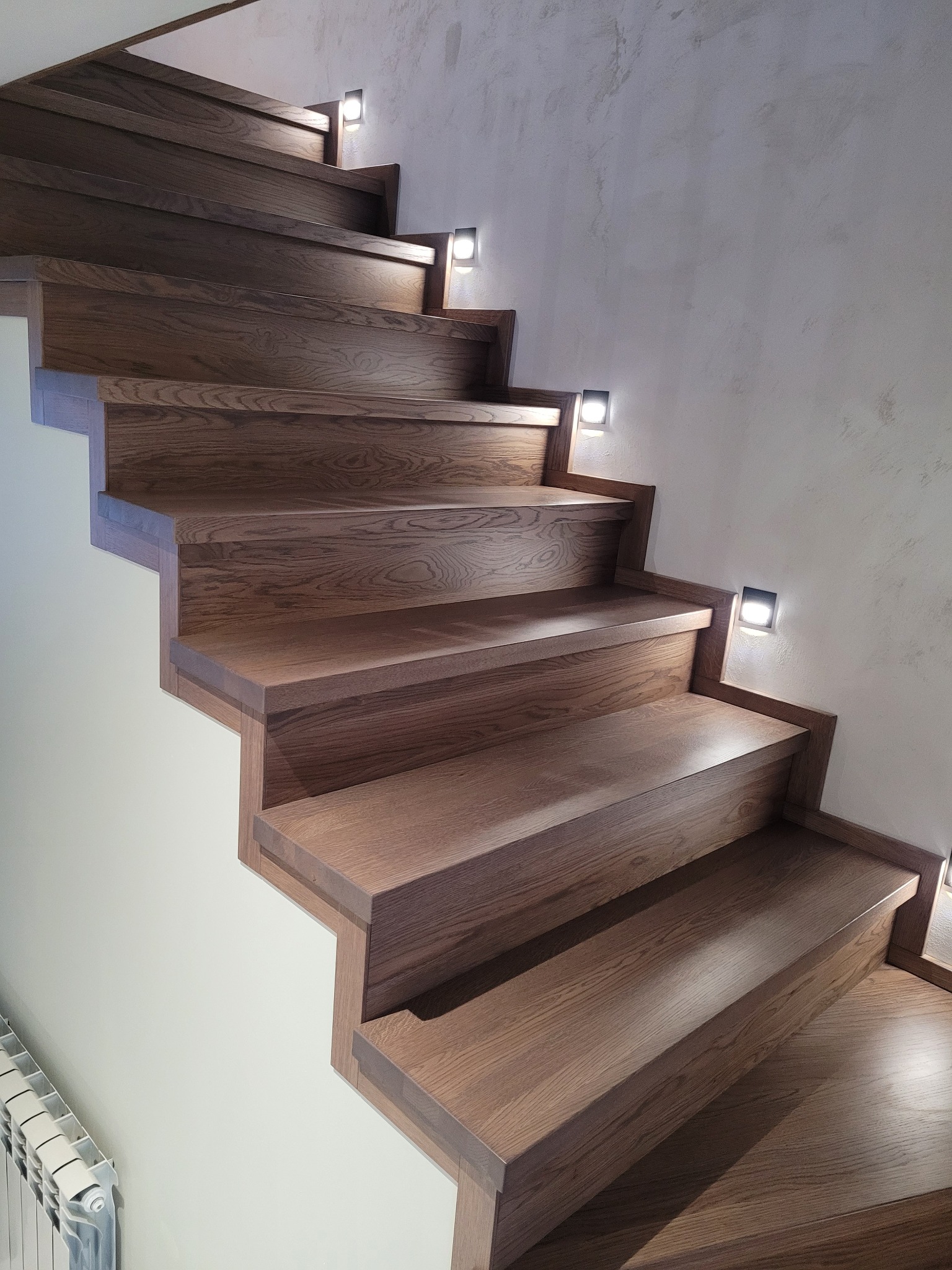 Nowoczesne schody z drewna o ciemnym odcieniu, podświetlane punktowymi lampkami, z widocznym fragmentem grzejnika. Minimalistyczny design i ciepłe oświetlenie.