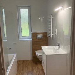 PG- firma remontowo - budowlana - Nowoczesna łazienka z drewnopodobnymi płytkami, białą armaturą i grzejnikiem drabinkowym. Okno z widokiem na zieleń.