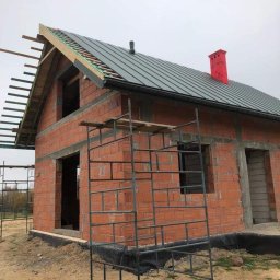 PG- firma remontowo - budowlana - Surowy, dwukondygnacyjny dom z czerwonej cegły w trakcie budowy. Na dachu szara blacha, czerwony komin. Widoczne rusztowania i elementy konstrukcji dachu.