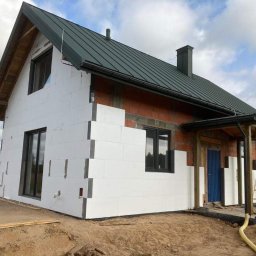 PG- firma remontowo - budowlana - Dom w trakcie ocieplania styropianem, widoczne fragmenty ściany z cegły. Dach pokryty blachą w kolorze ciemnej zieleni. Okna czarne.