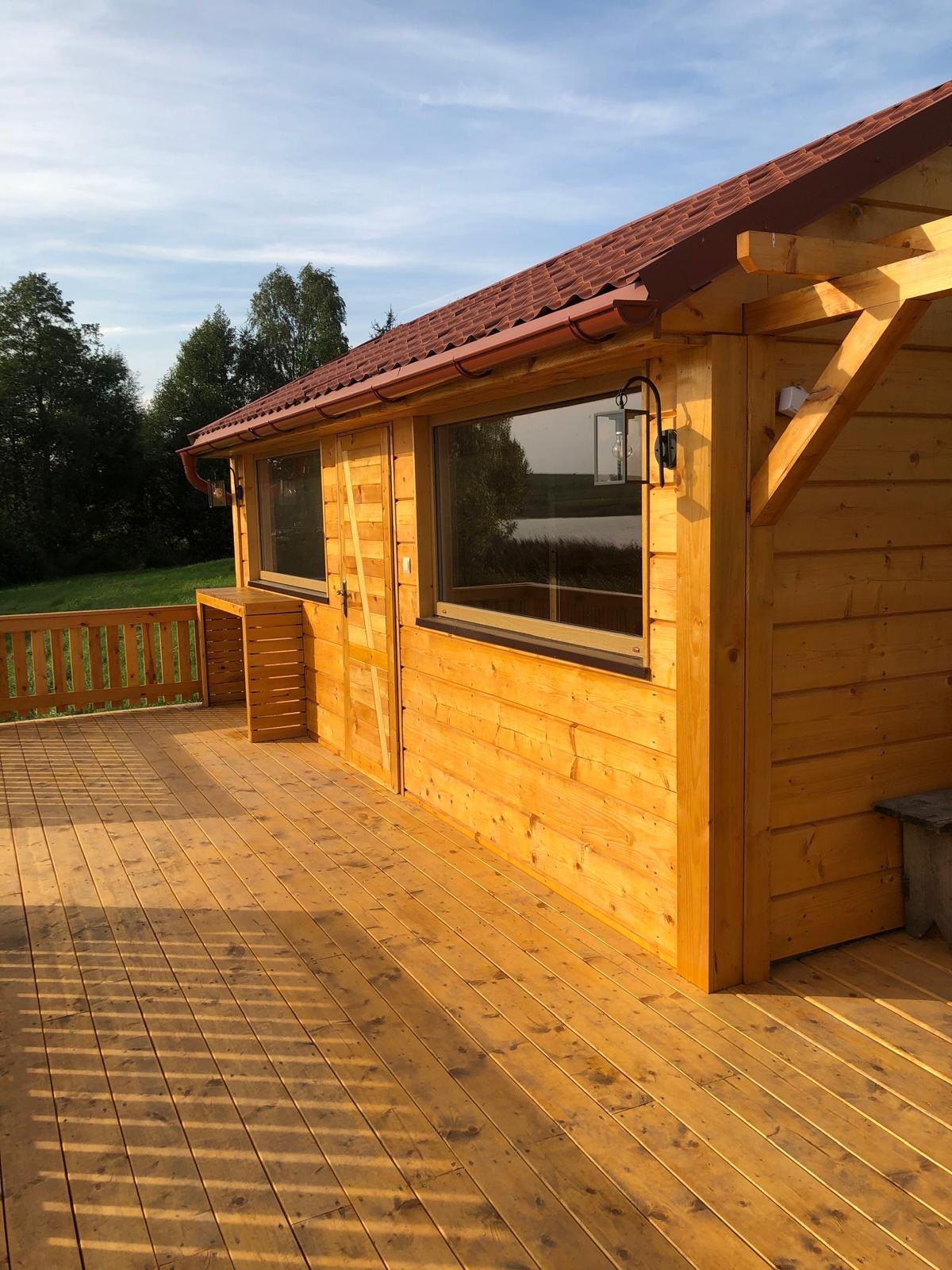 Drewniany domek letniskowy z tarasem, widok z zewnątrz. Czerwona dachówka, drewniane balustrady tarasu. Słoneczny dzień, zielone drzewa w tle.