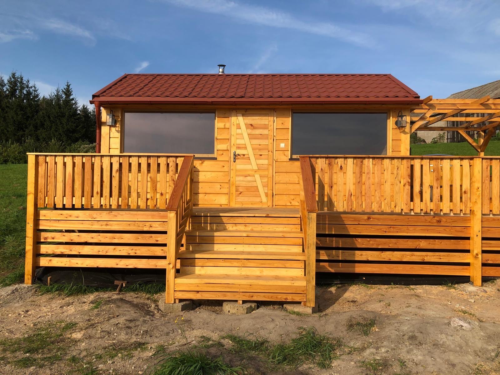 Drewniany domek letniskowy z tarasem i schodami, kryty czerwoną dachówką, widziany w słoneczny dzień. Elementy wykończeniowe z surowego drewna.